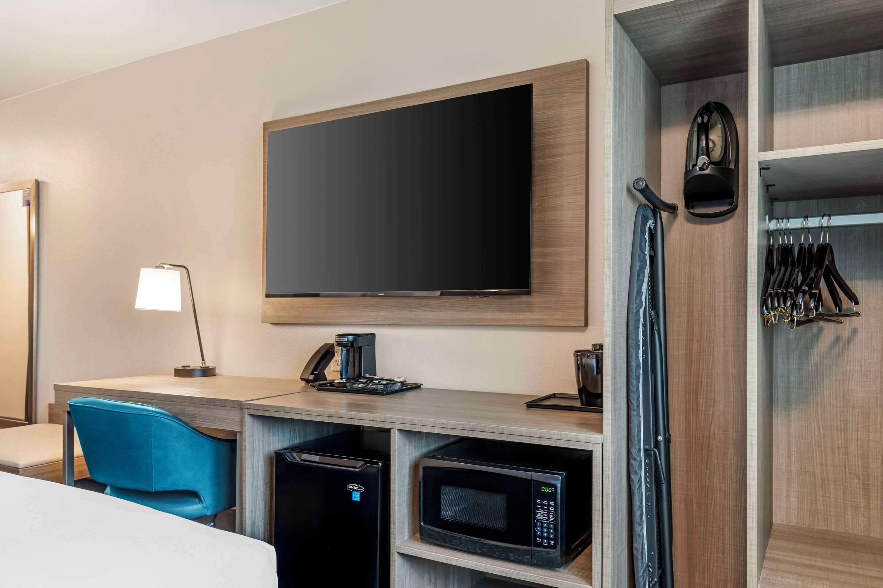 A hotel room with a flat-screen TV, a desk with a coffee maker, a microwave, a mini fridge, a blue chair, and a coat rack with hanging items.