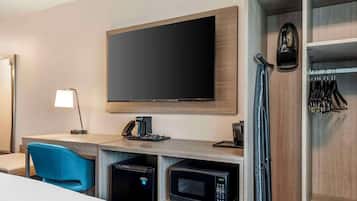 Habitación de hotel con televisión de pantalla plana, escritorio con cafetera, microondas, refrigerador pequeño, una silla azul y perchero con objetos colgados.