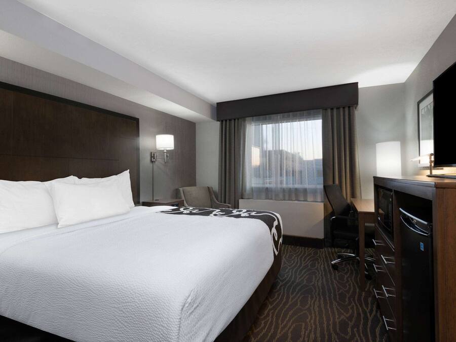 Quarto de hotel com uma cama grande, uma mesa com televisão de tela plana, uma cadeira, abajur e uma janela com cortinas.