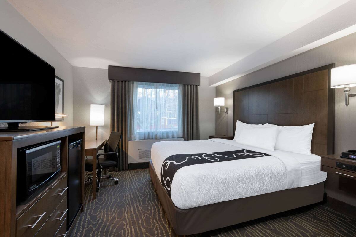 Habitación de hotel con una cama grande, un escritorio con silla, televisión y ventana con cortinas.
