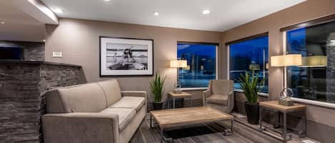 A modern living room with a stone fireplace, a sofa, a coffee table, and two armchairs. There are two windows with city views at night.