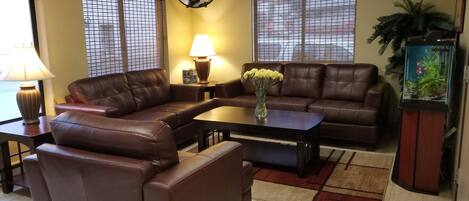 A living room with a brown leather sofa, a coffee table, and a lamp. There is a window with blinds and a potted plant.
