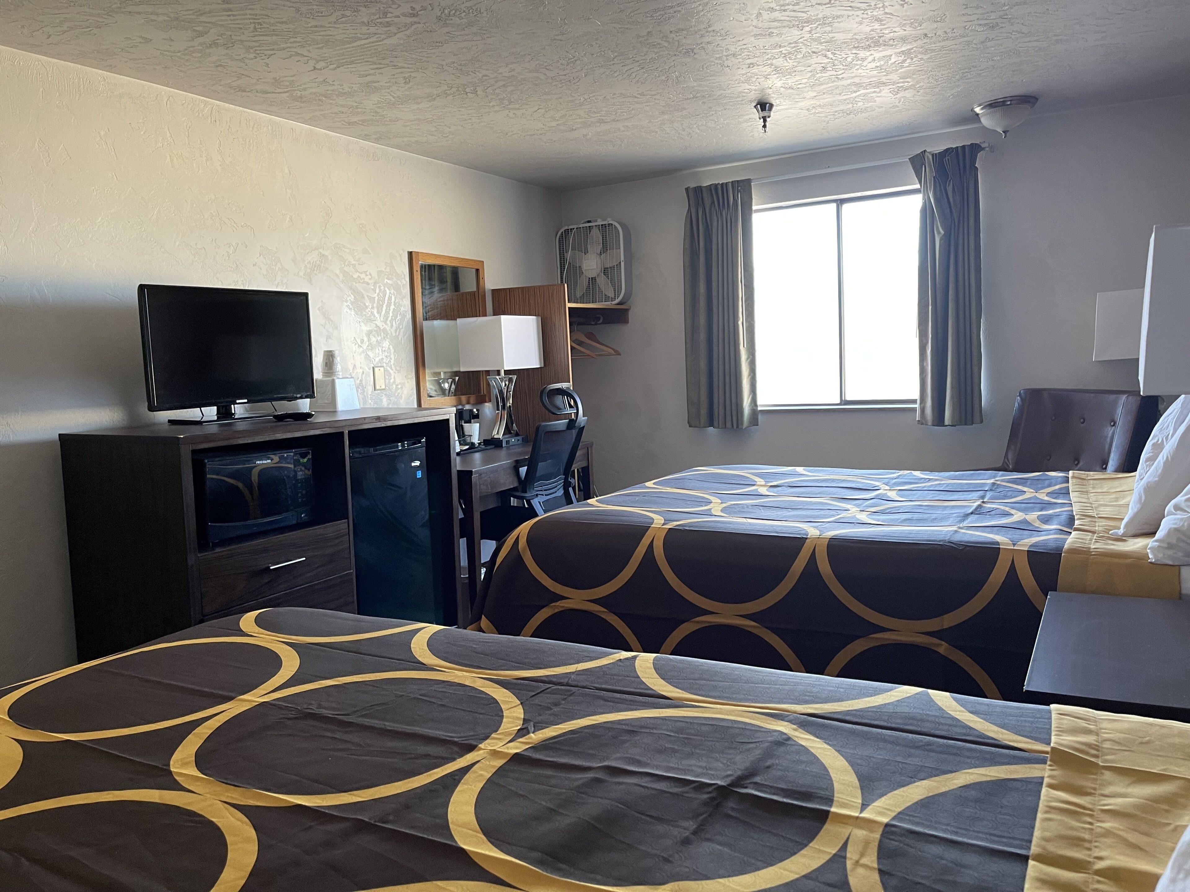 Ein Hotelzimmer mit zwei Betten, einem Schreibtisch mit Computer, einem Fernseher und einem Fenster mit Vorhängen.
