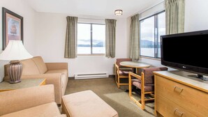 Ein Hotelzimmer mit einer Couch, einem kleinen Tisch mit Lampe, einem Fernseher, einem Sessel und einem Schreibtisch mit Schubladen.