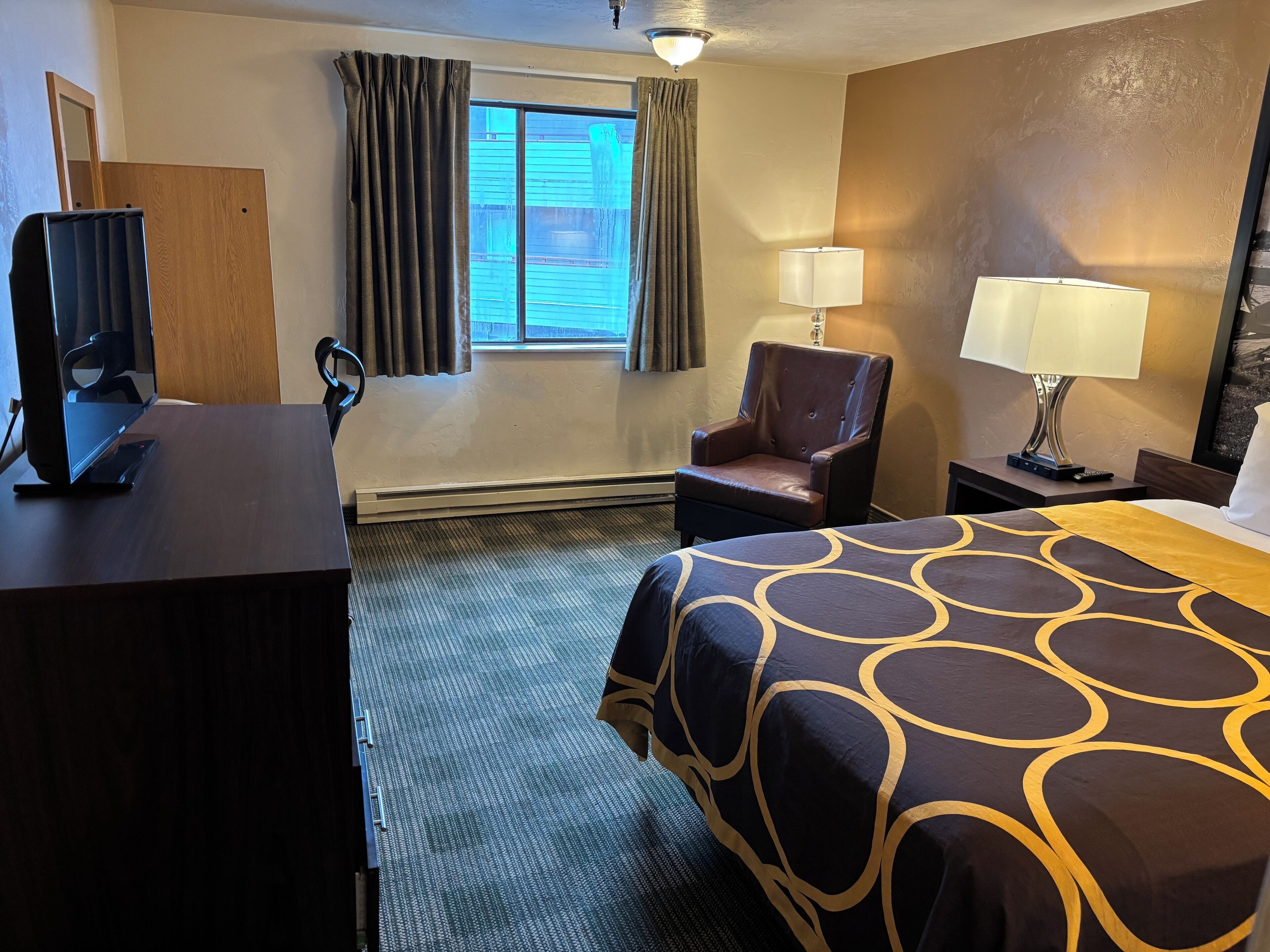 Ett hotellrum med en säng, ett skrivbord, en stol, en tv och ett fönster med gardiner.