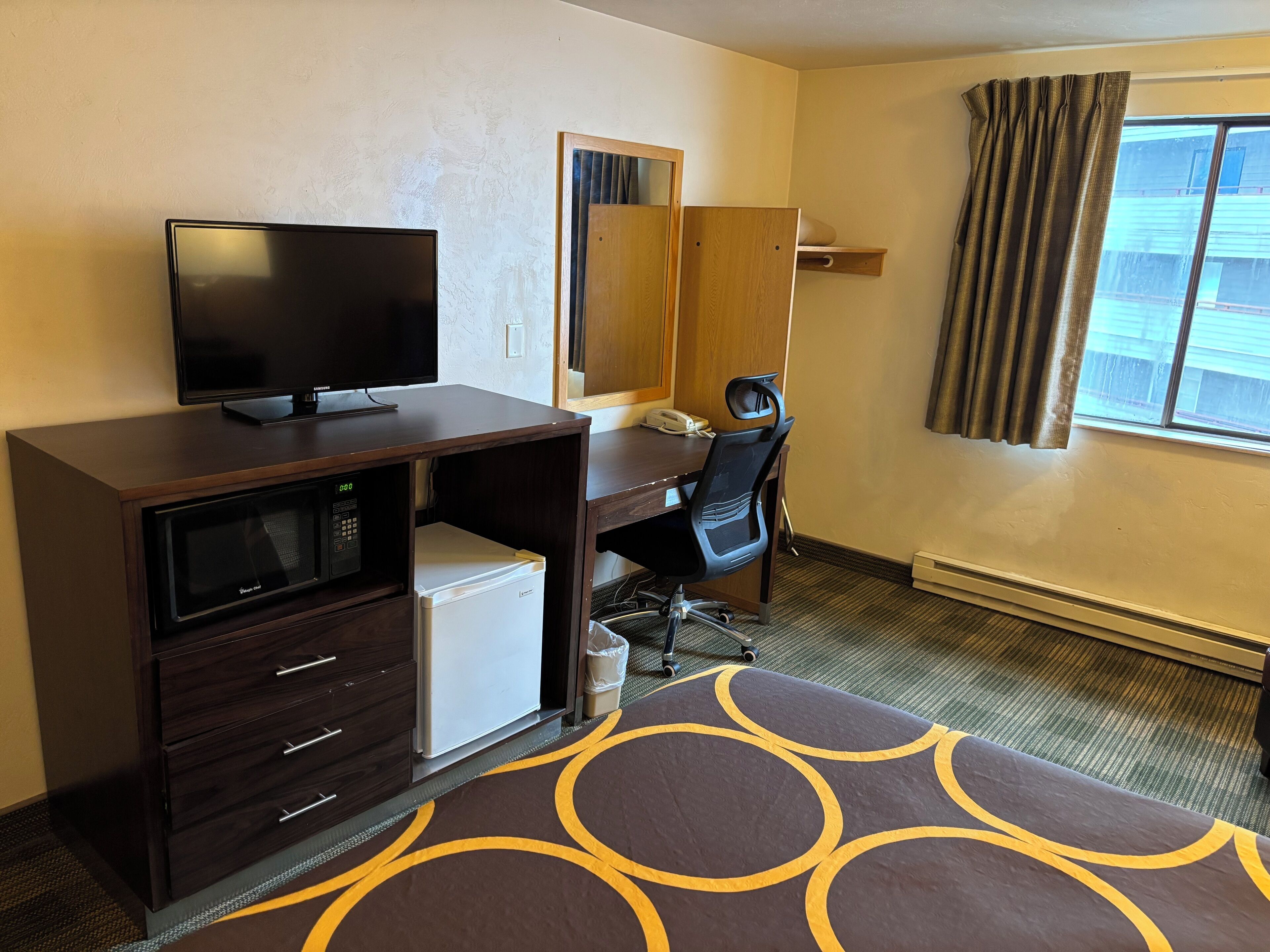 A hotel room with a desk, chair, television, microwave, refrigerator, and a window with curtains.