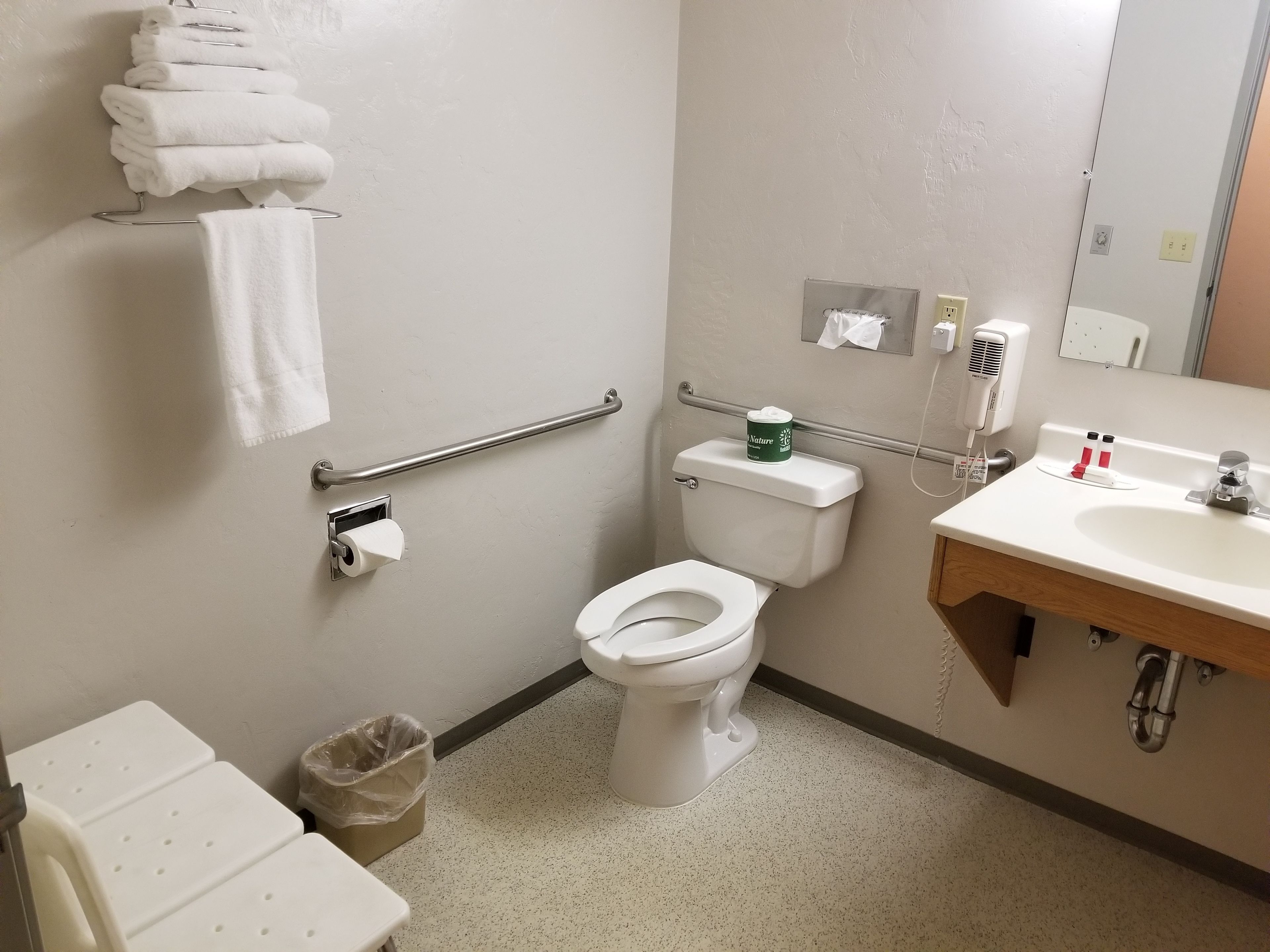 A bathroom with a toilet, sink, handrails, and a trash bin.