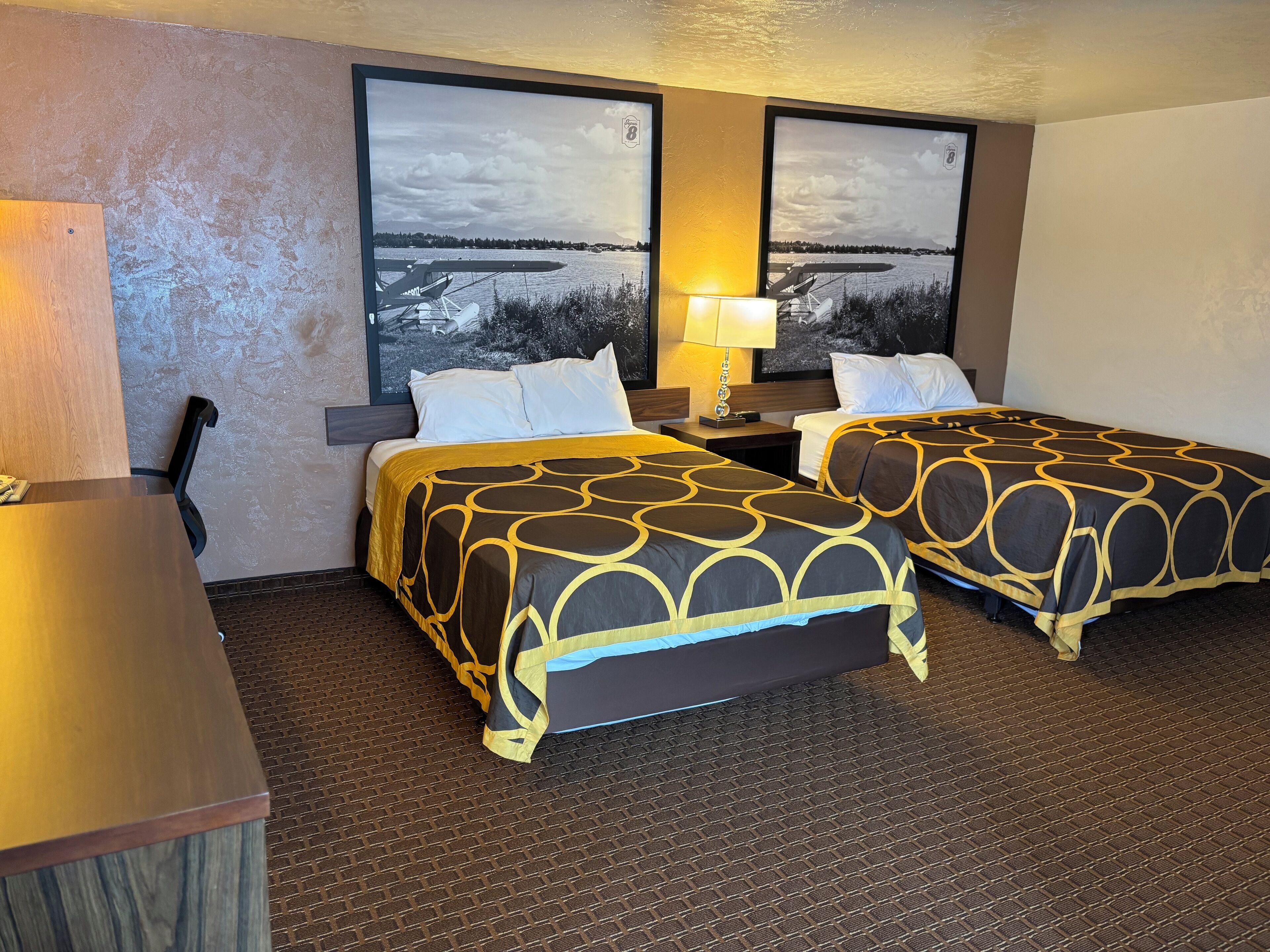 Quarto de hotel com duas camas, uma escrivaninha e uma janela grande com vista para a água e um céu nublado.