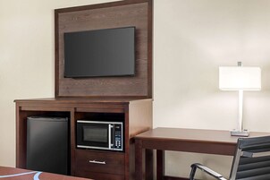 Habitación de hotel con un escritorio de madera, un televisor de pantalla plana montado encima, un microondas, un cubo de basura y una silla.