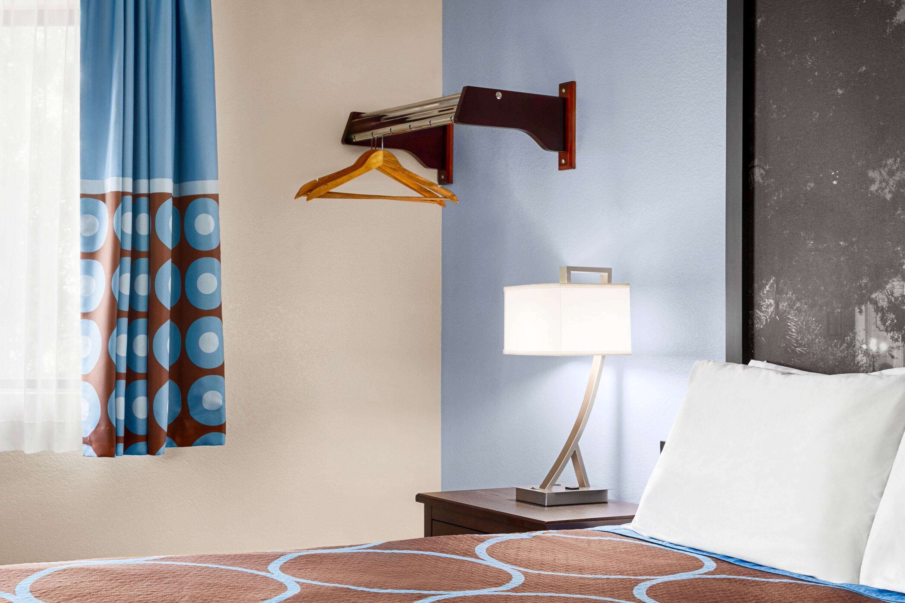 Una habitación de hotel con una cama, una mesita de noche, una lámpara, un perchero en la pared y una cortina con un diseño circular.