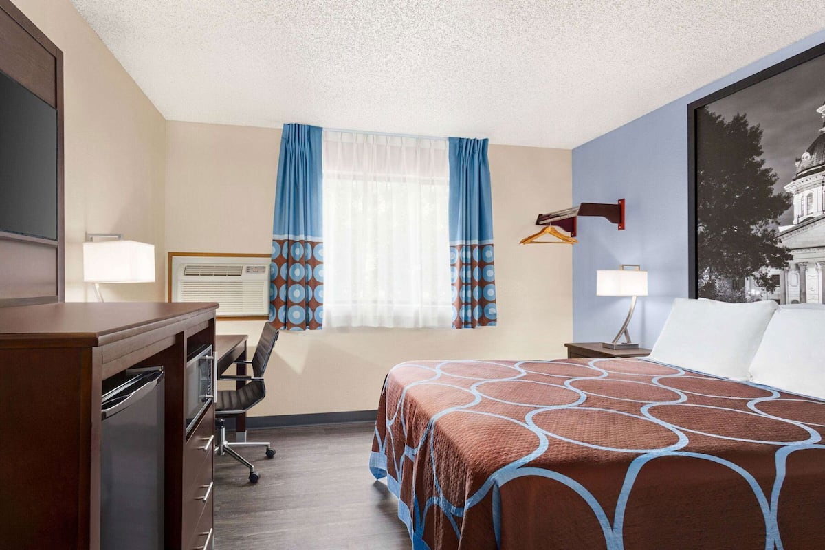 A hotel room with a bed, desk, chair, and a large window with curtains.