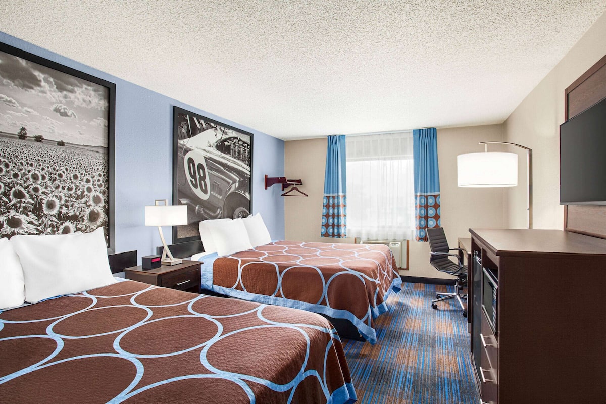 A hotel room with two beds, a desk, a TV, and a large window with curtains.