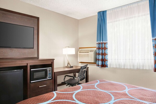 A hotel room with a bed, a desk with a chair, a microwave, a refrigerator, a TV, and a window with curtains.
