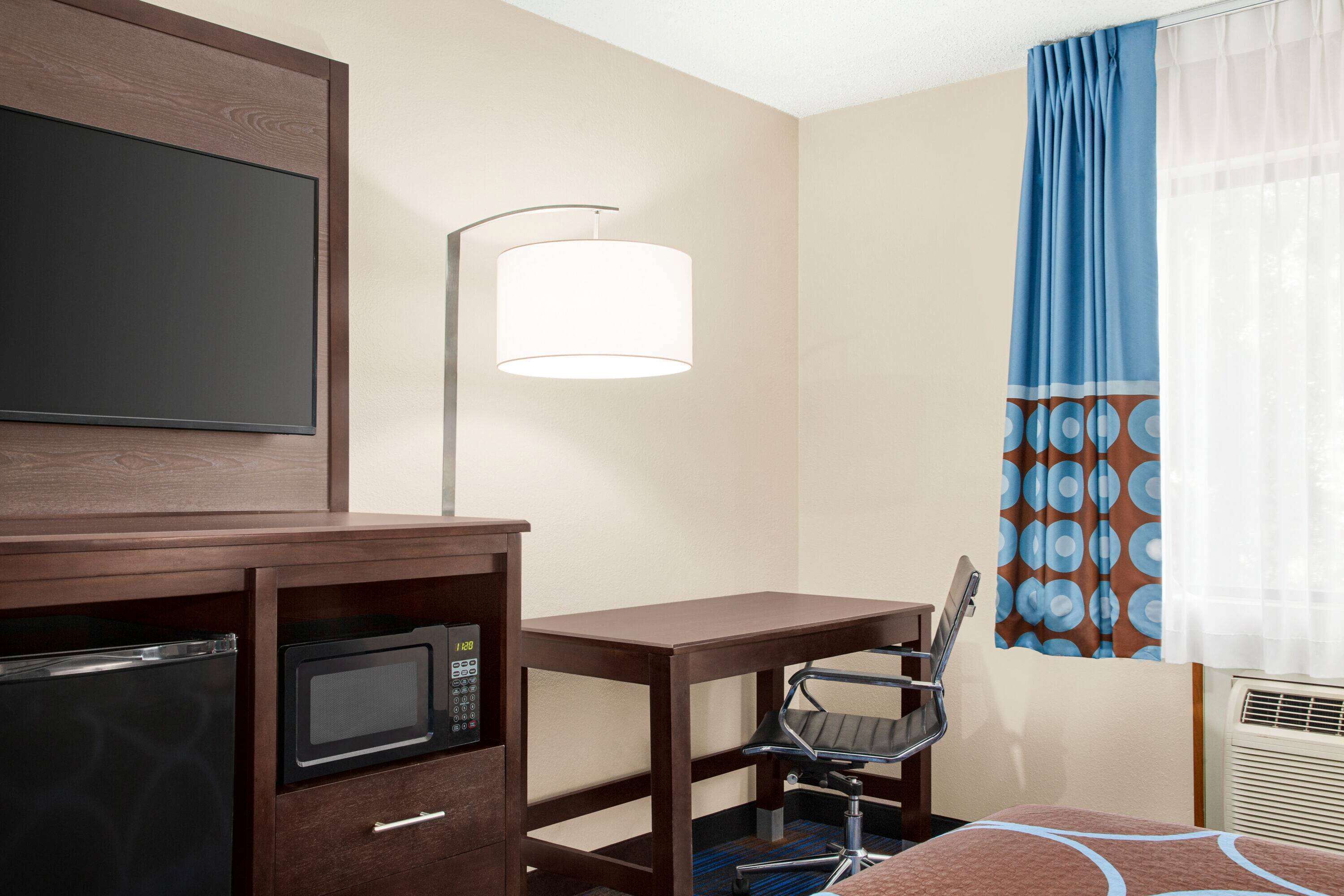 A hotel room with a desk, chair, microwave, and a TV mounted on the wall.