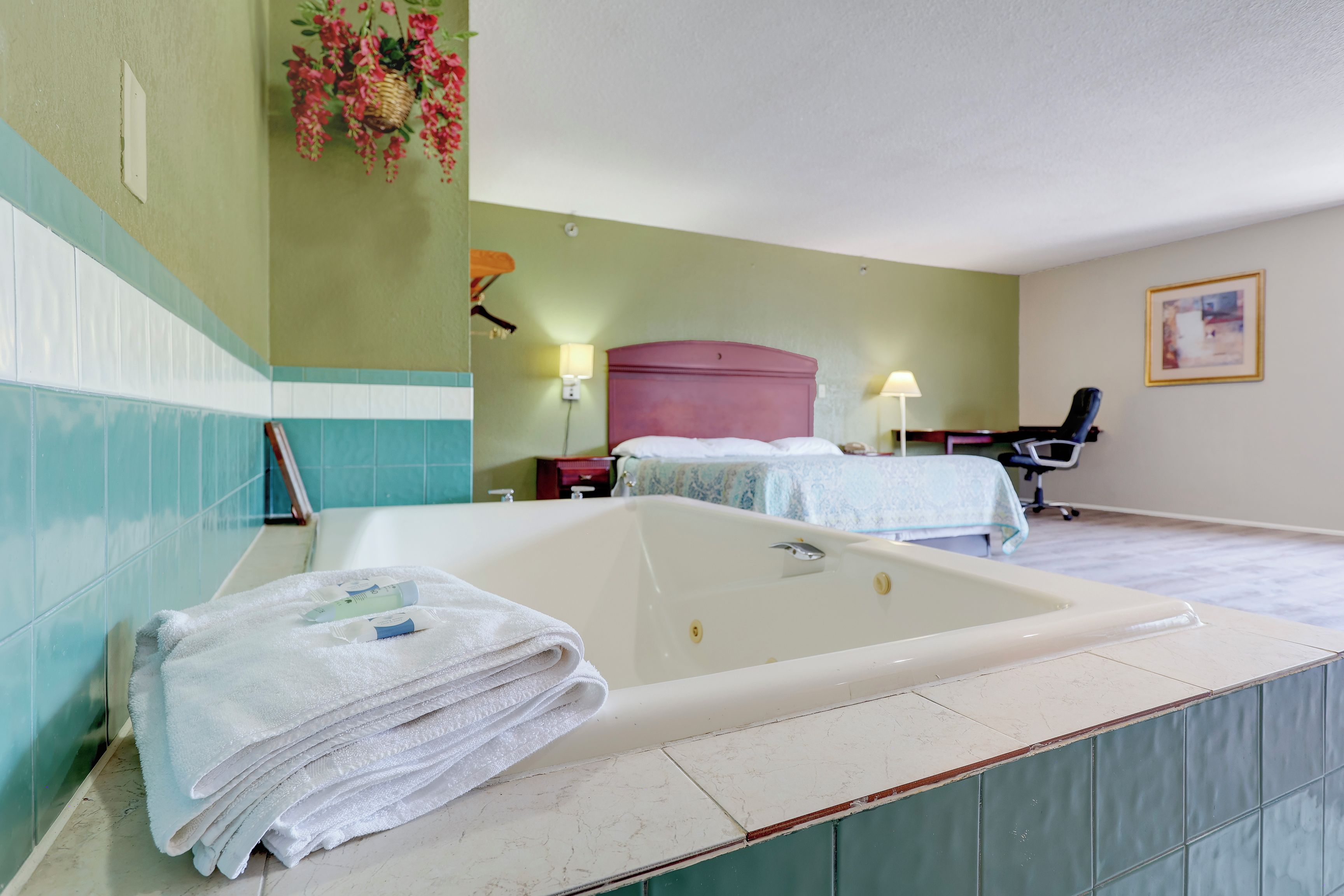 A hotel room with a large bathtub, a bed with a headboard, a desk with a chair, and a wall-mounted lamp.