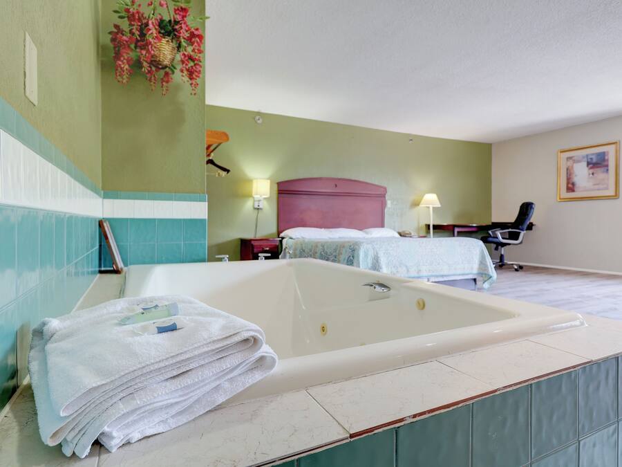 A hotel room with a large bathtub, a bed with a headboard, a desk with a chair, and a wall-mounted lamp.