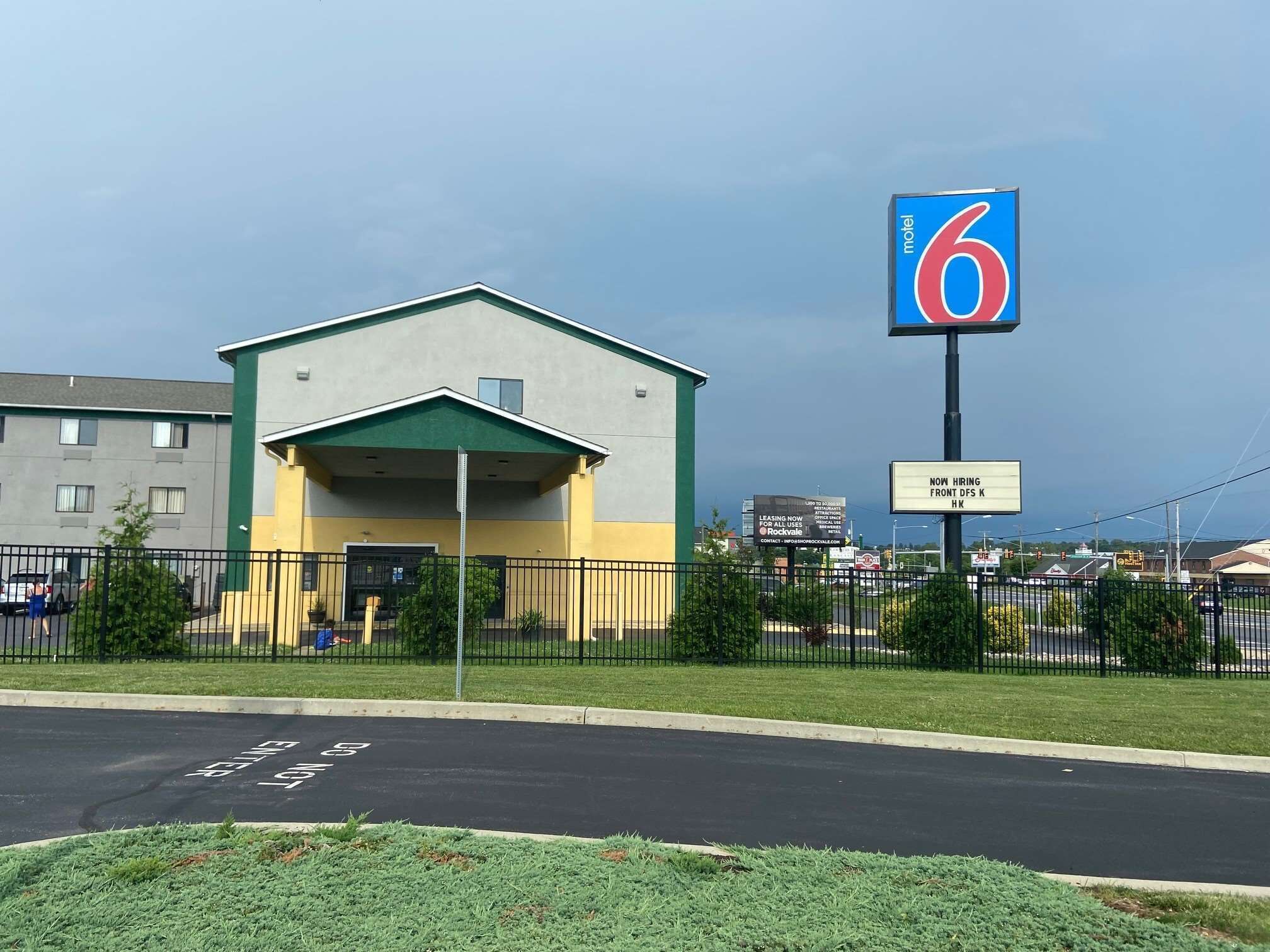 Un motel con facciata verde e gialla, un parcheggio e un grande cartello che indica '6' e 'ASSUNZIONI IN CORSO'.