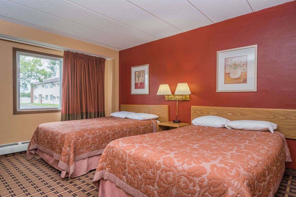 Une chambre d’hôtel avec deux lits, une fenêtre avec des rideaux, une tête de lit et deux lampes fixées au mur.