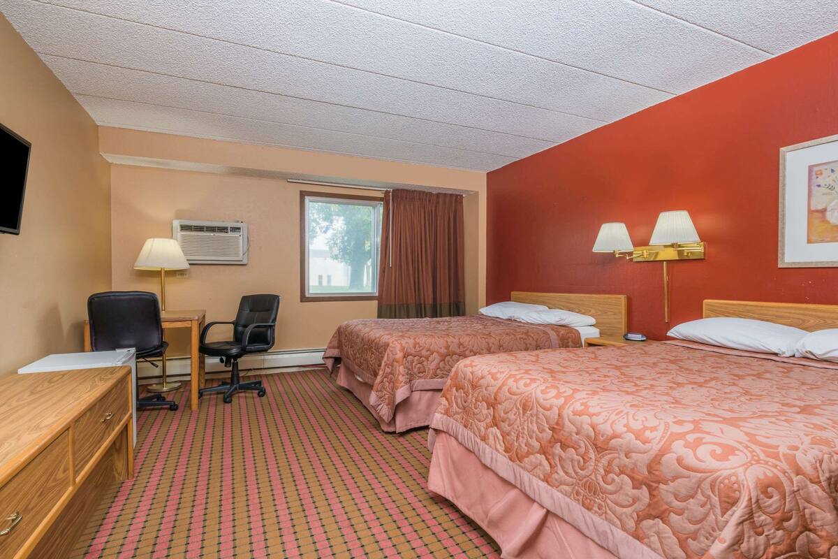 Une chambre d’hôtel avec deux lits, un bureau, une chaise, une lampe et une fenêtre avec des rideaux.