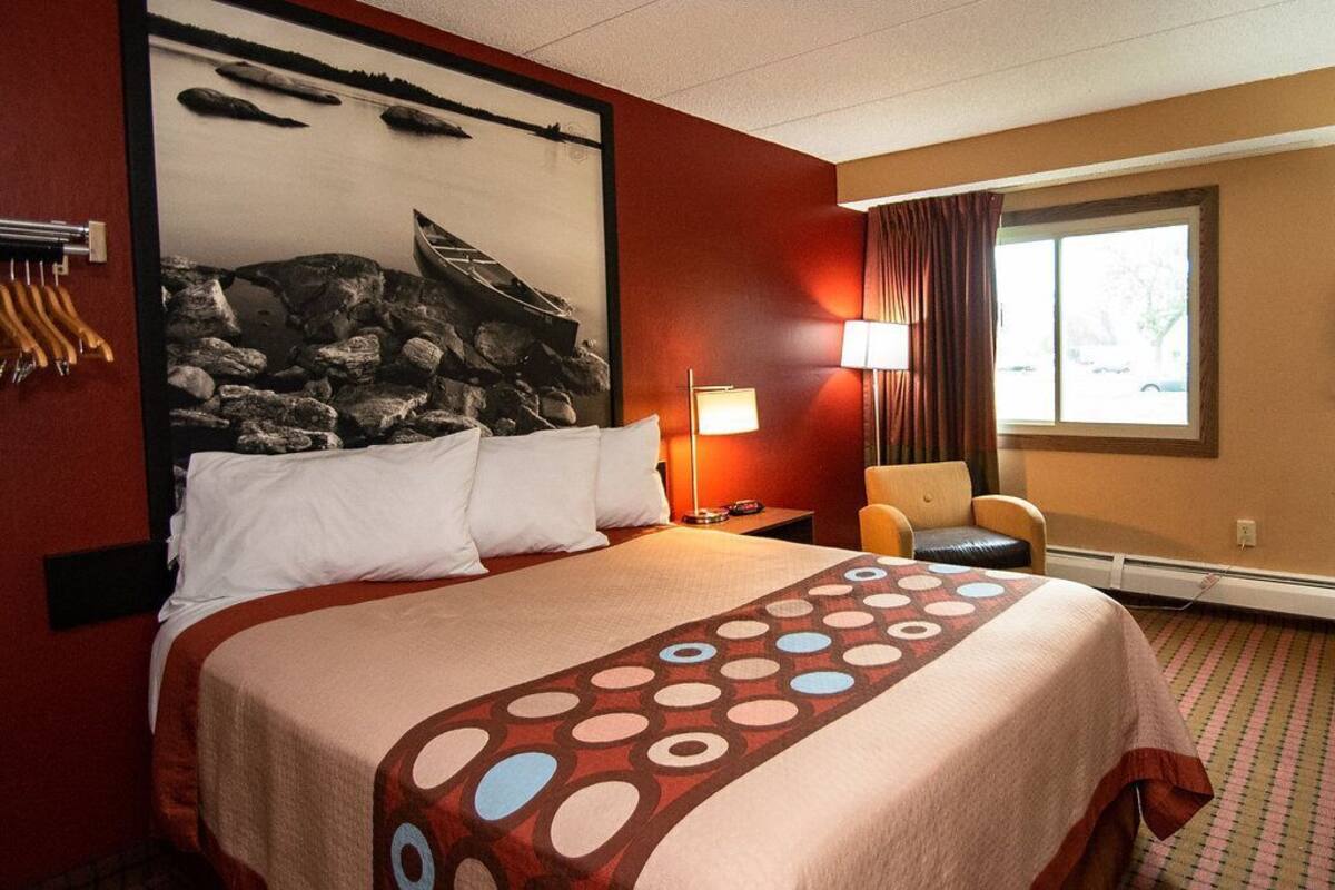 Une chambre d’hôtel avec un lit, une chaise, une lampe, une fenêtre et un mur orné d’un tableau.
