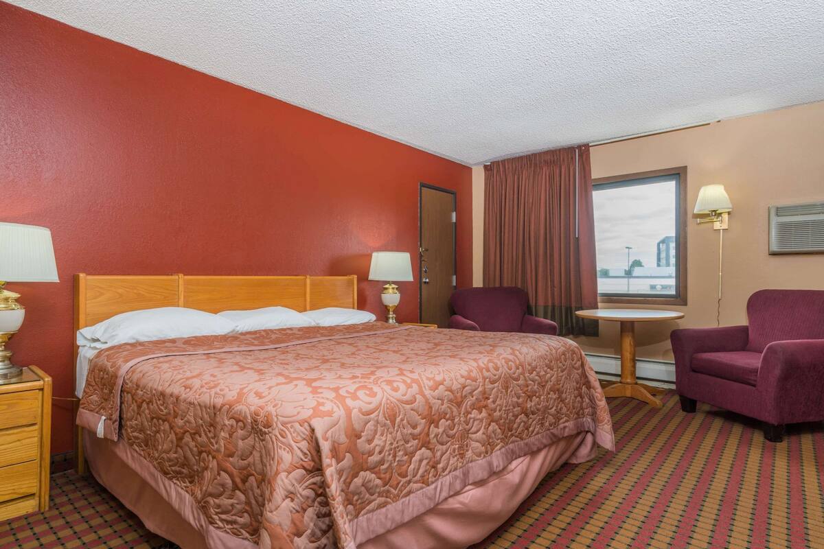 Une chambre d’hôtel avec un grand lit, une tête de lit en bois, une petite table, une chaise et une fenêtre avec des rideaux.