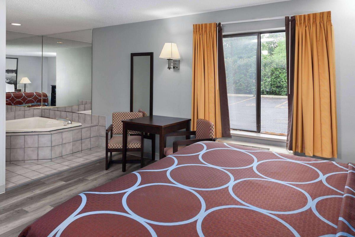 A hotel room with a large bed, a desk, a chair, a window with curtains, a bathtub, and a mirror.