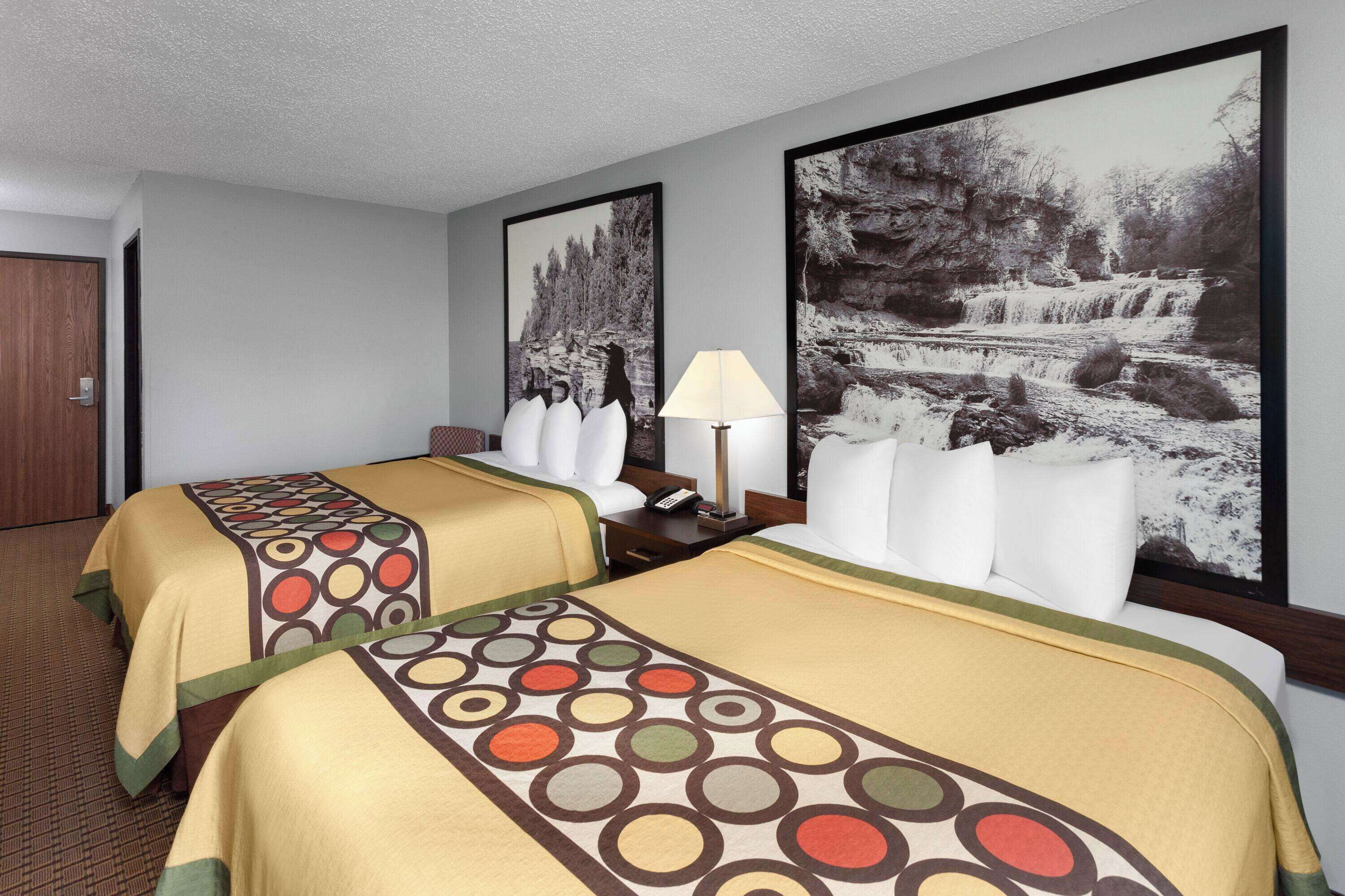 A hotel room with two beds, a nightstand, a lamp, and large framed pictures of a waterfall and a forest.