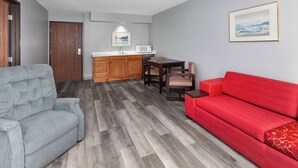 A compact living space with a grey sofa, a red sofa, a wooden dining table with chairs, a kitchen area with a microwave, and a framed picture on the wall. - Super 8 by Wyndham Sun Prairie/Madison E (Sun Prairie)