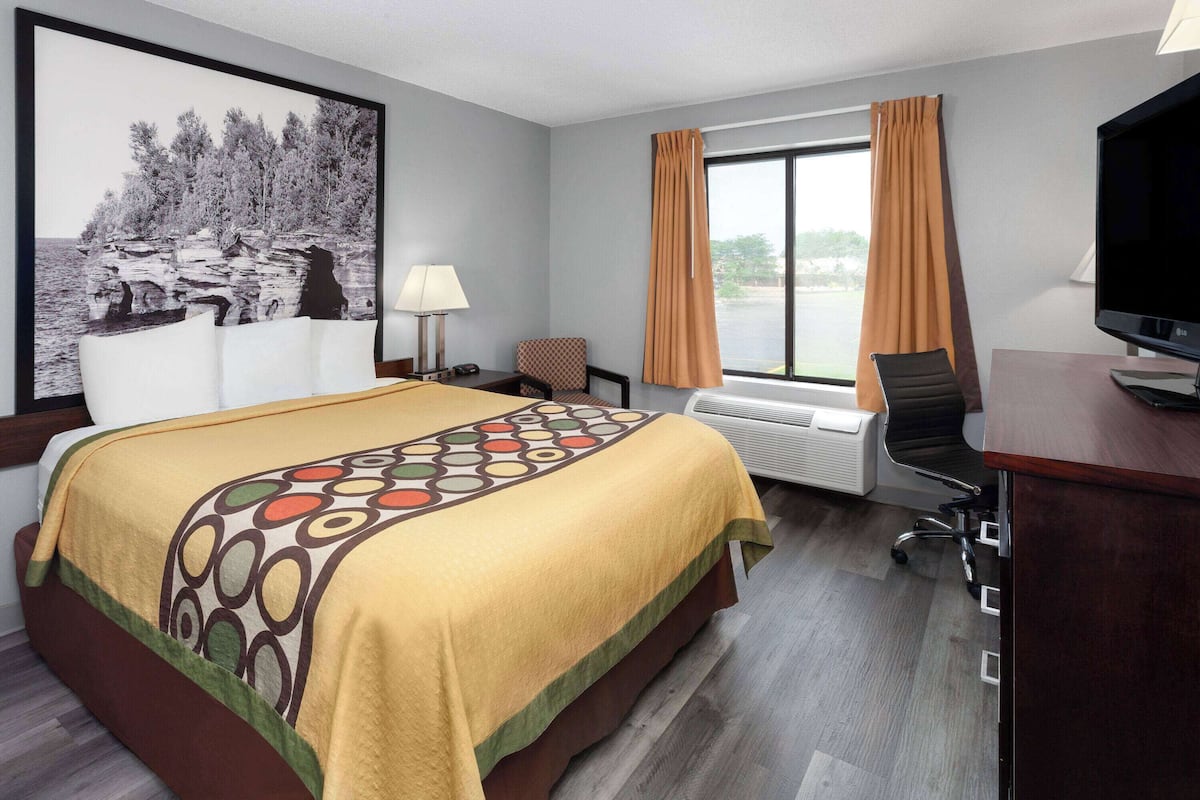 A hotel room with a bed, a desk with a TV, a chair, a window with curtains, and a painting on the wall.
