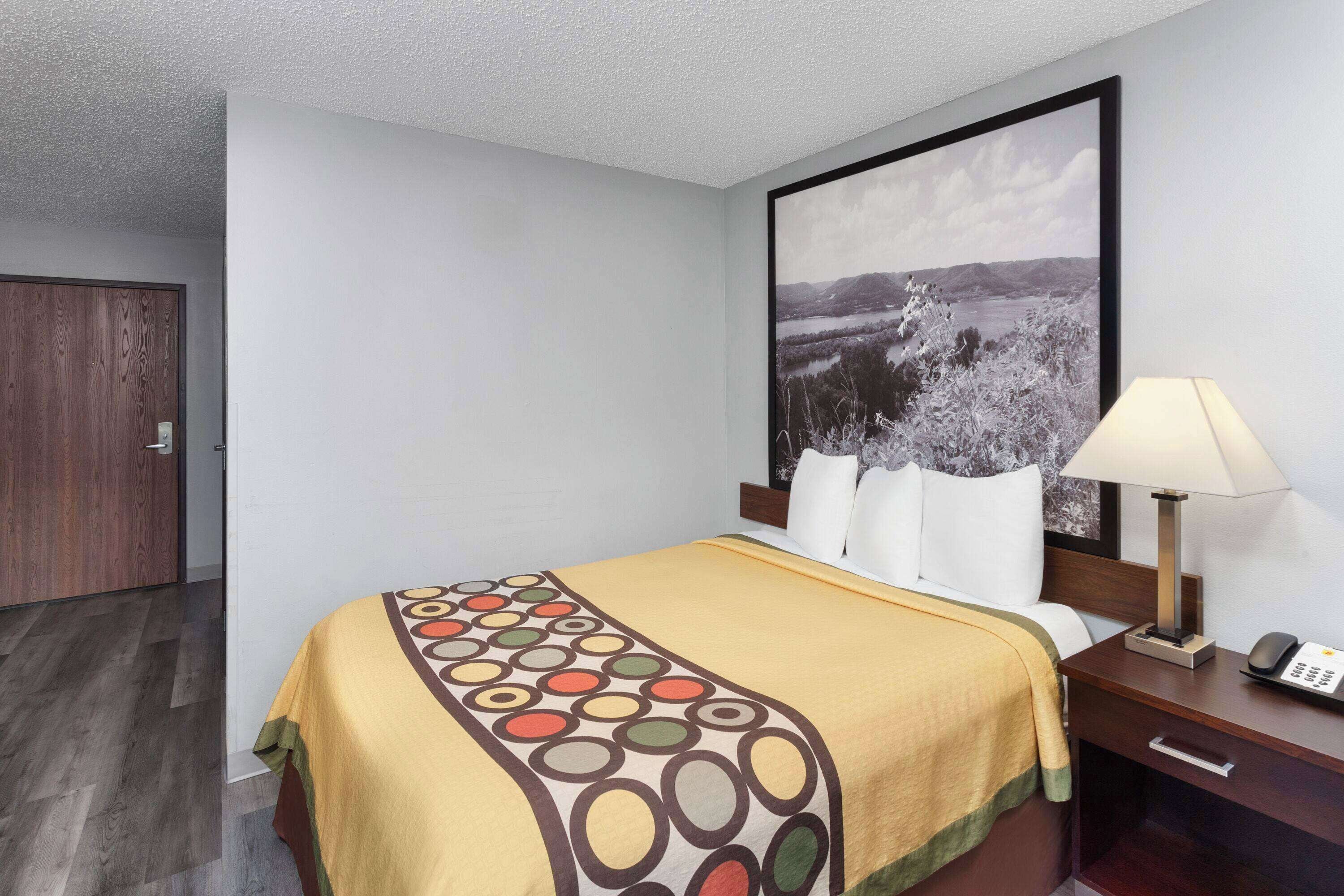 A hotel room with a bed, a nightstand with a lamp, a telephone, and a framed picture on the wall.