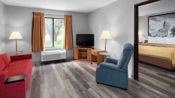 Une chambre d’hôtel avec un canapé rouge, un fauteuil bleu, une télévision, un bureau avec une lampe et un tableau représentant un bâtiment du Capitole.