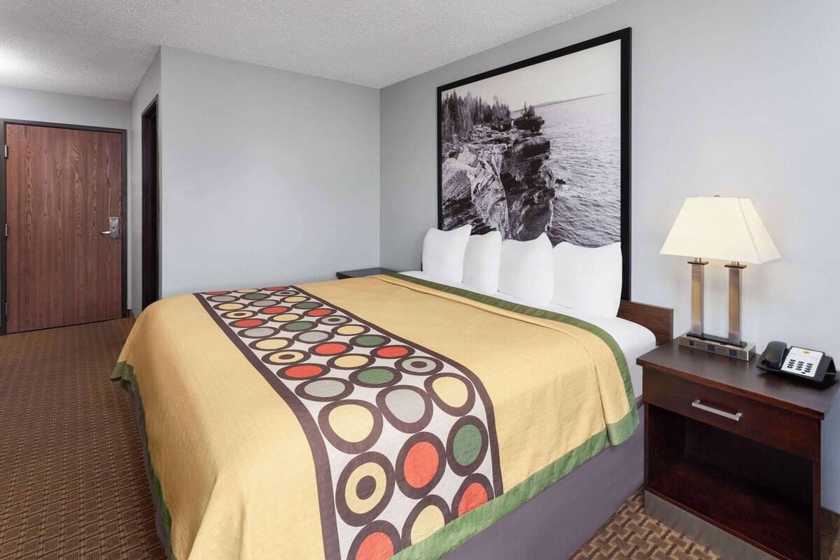 A hotel room with a bed, a nightstand with a lamp, a framed picture on the wall, and a door.