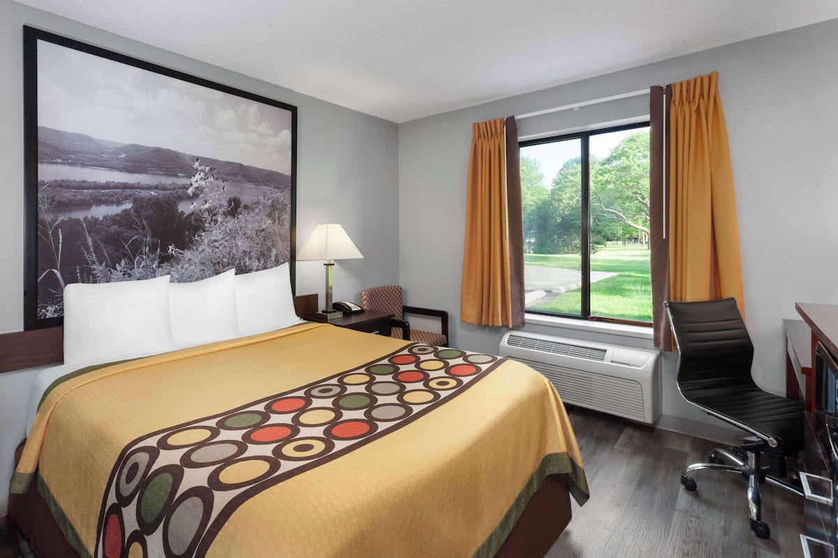 A hotel room with a large bed, a desk with a chair, a window with curtains, and a painting on the wall.