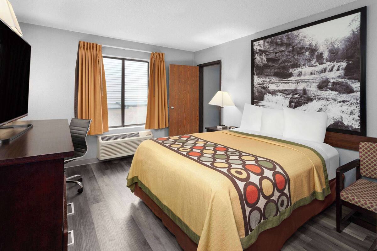 A hotel room with a bed, a desk, a chair, a window with curtains, a wall-mounted TV, and a large framed picture.