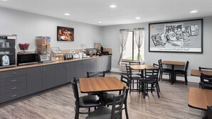 A modern dining area with a kitchen counter, microwave, and various food items. There are wooden tables and chairs, and a large framed picture on the wall. - Super 8 by Wyndham Sun Prairie/Madison E (Sun Prairie)