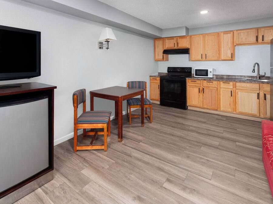 A compact living space with a TV, dining table, chairs, a kitchen area with wooden cabinets, a microwave, and a refrigerator.