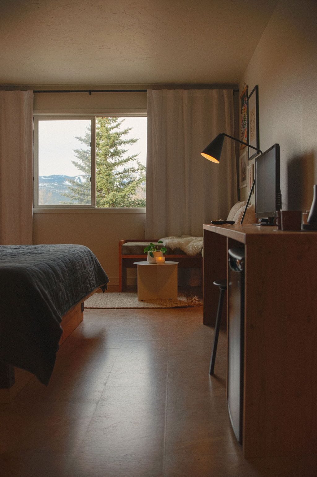 Et soveværelse med seng, skrivebord, stol, lampe og et vindue med udsigt til træer og bjerge.