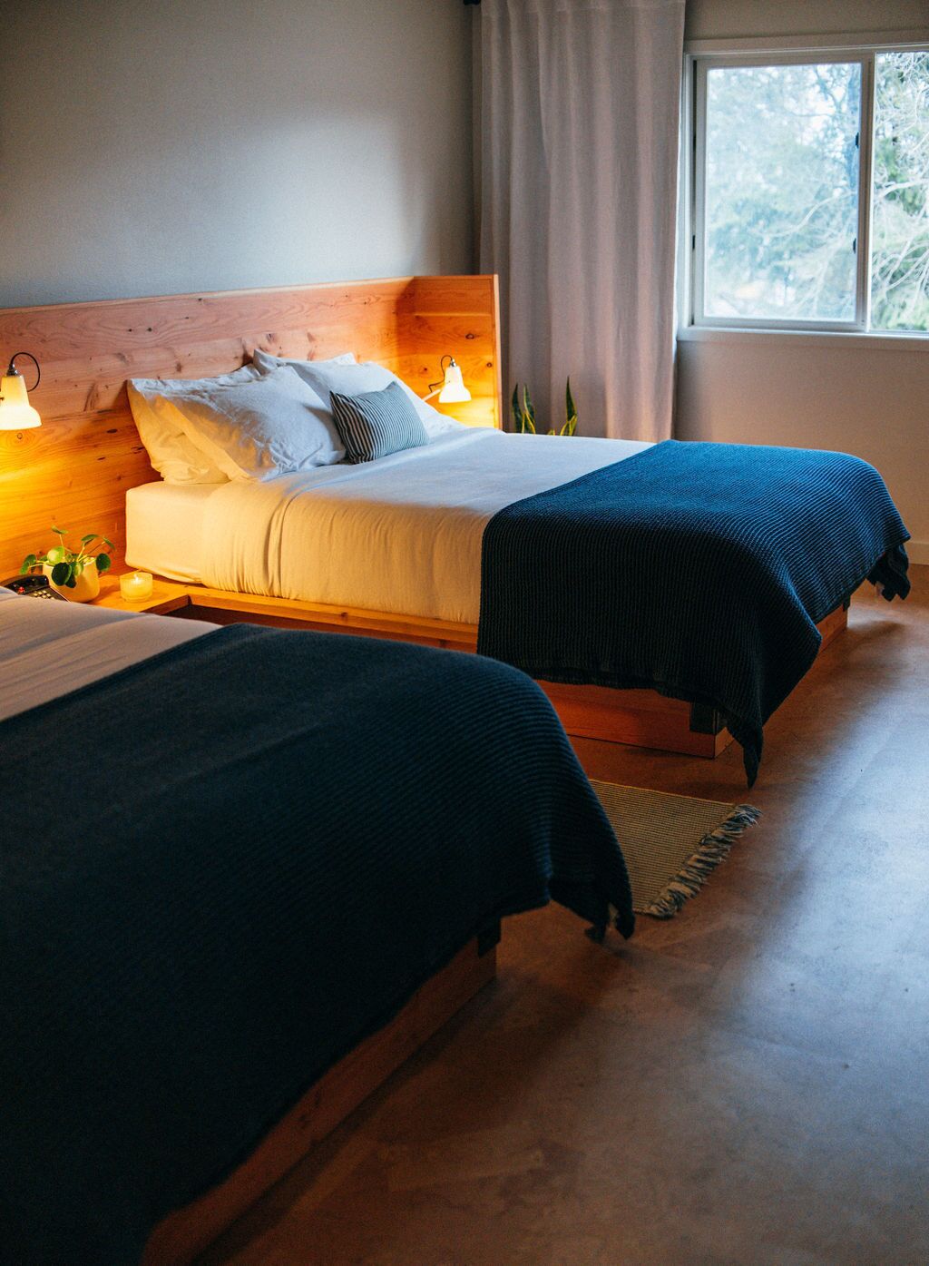 Quarto com duas camas, cabeceira de madeira, luminárias de cabeceira, janela com cortinas e vista para árvores.