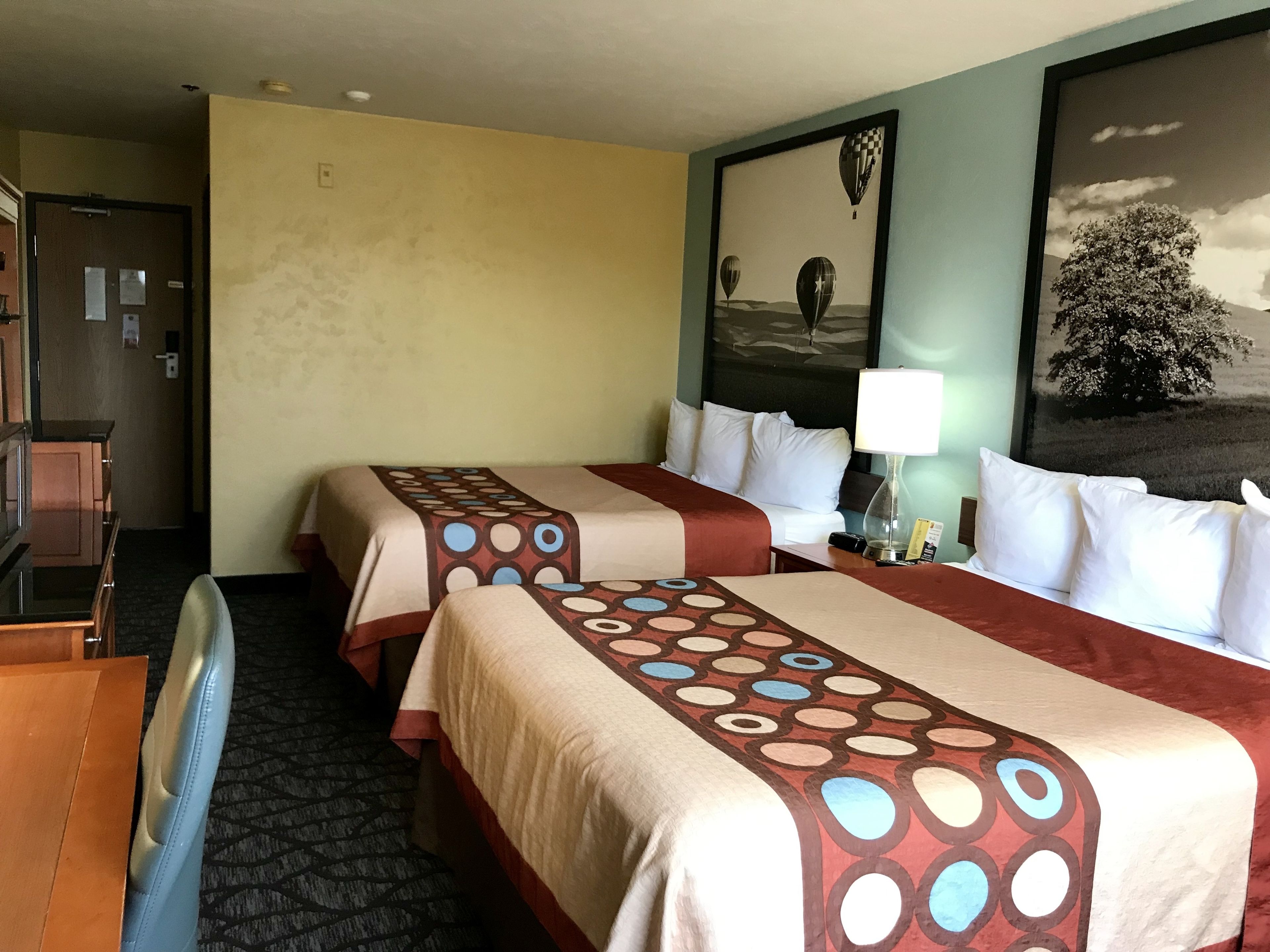 A hotel room with two beds, a desk, a lamp, and a painting on the wall.