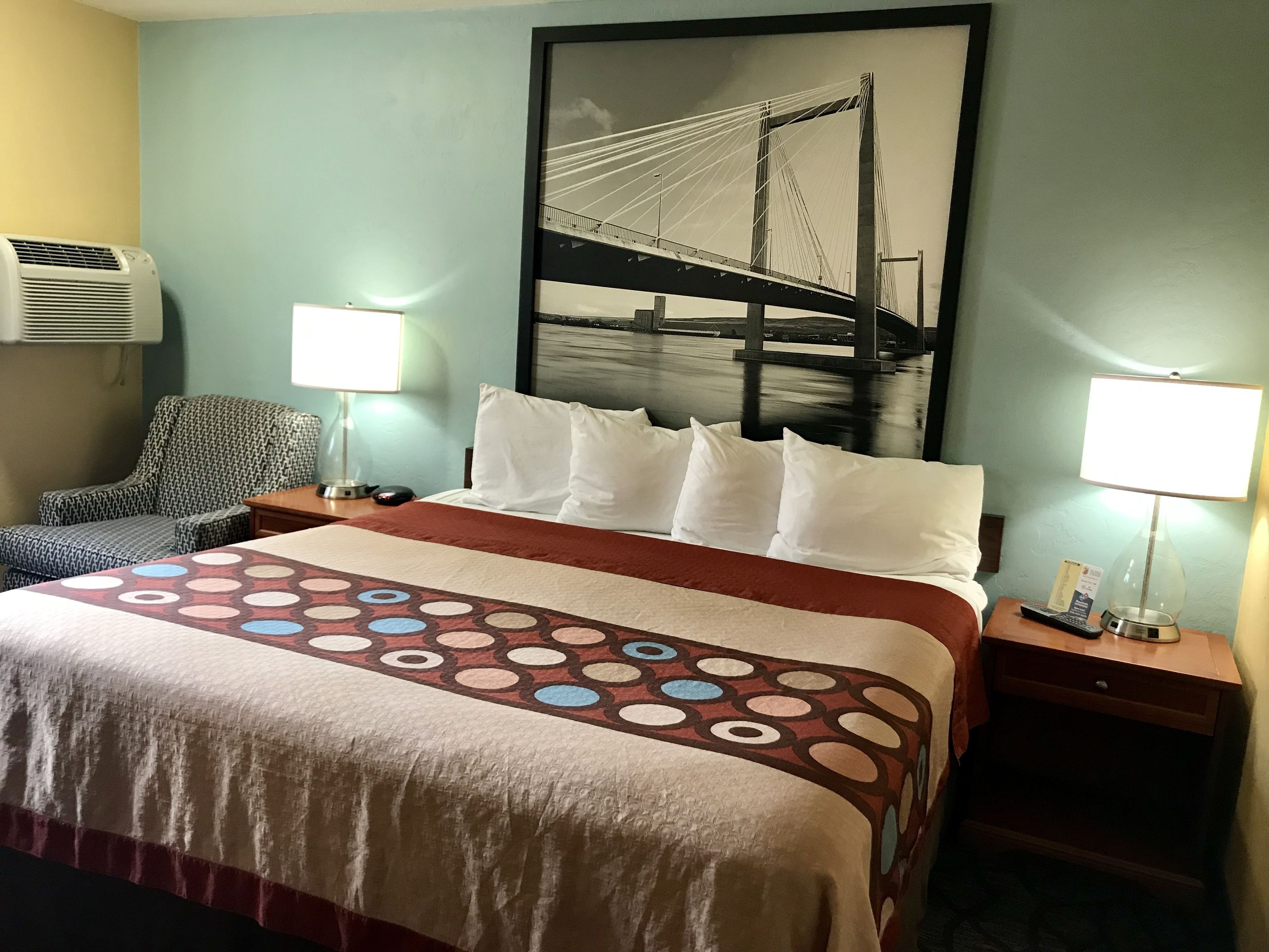 Quarto de hotel com uma cama, duas mesas de cabeceira, uma cadeira, ar condicionado fixado na parede e um quadro grande de uma ponte.