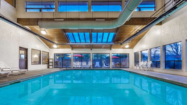 Ein Innenpool mit klarem, blauem Wasser, umgeben von weißen Liegestühlen und Sonnenschirmen, mit großen Fenstern und hoher Decke.
