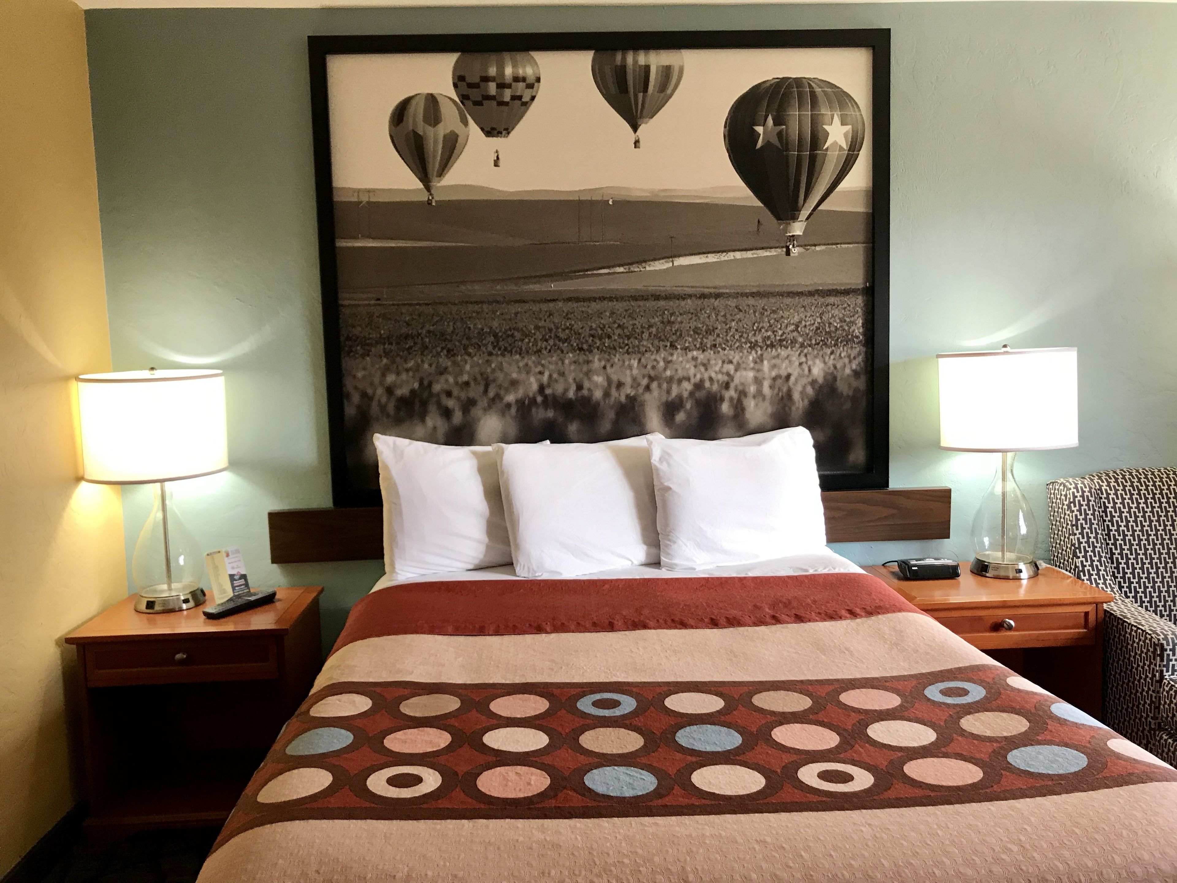 Una habitación de hotel con una cama, dos lámparas de noche, un cuadro enmarco con globos aerostáticos y una colcha estampada.