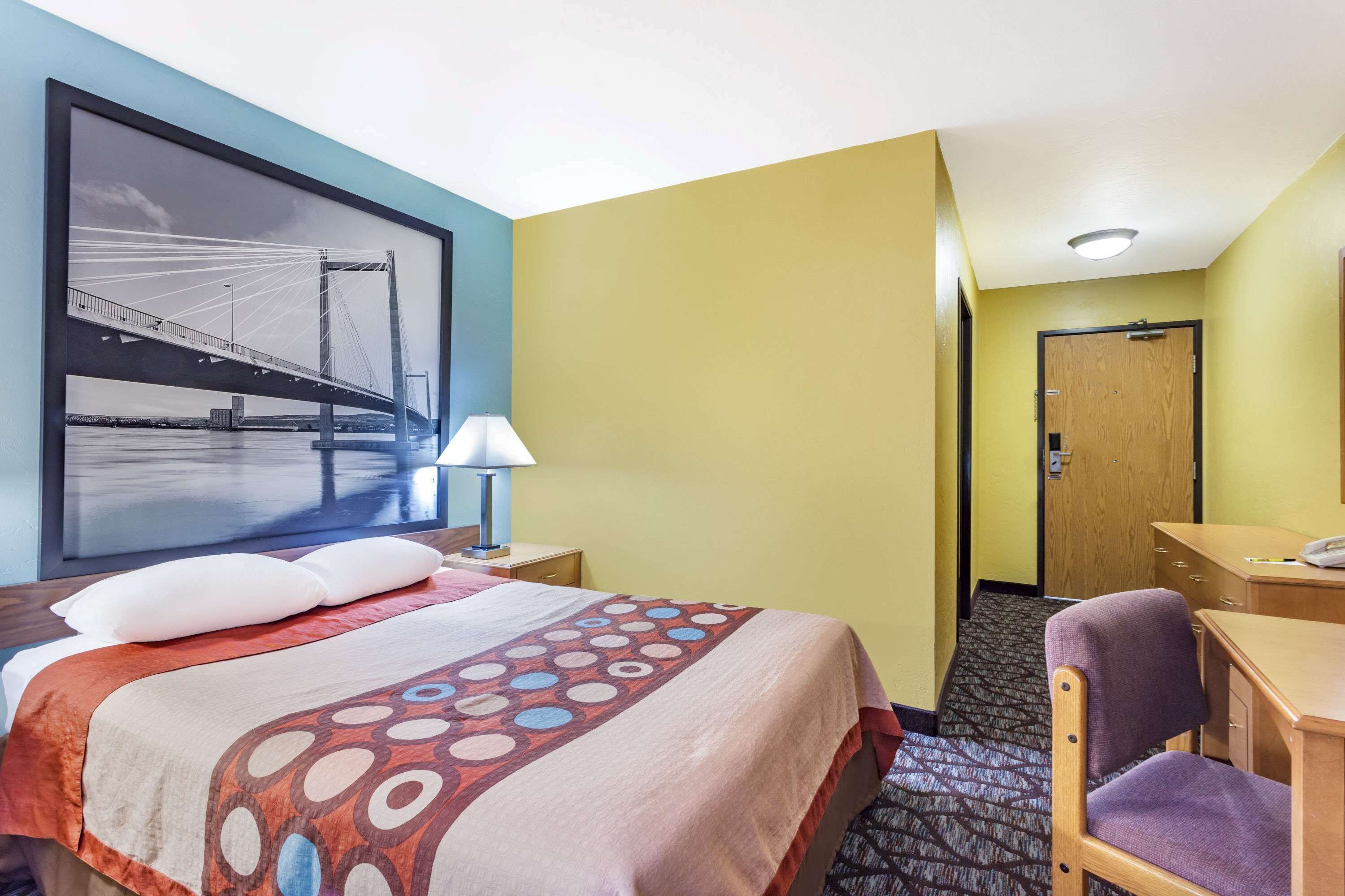 A hotel room with a bed, a desk, a chair, a lamp, and a framed picture of a bridge.