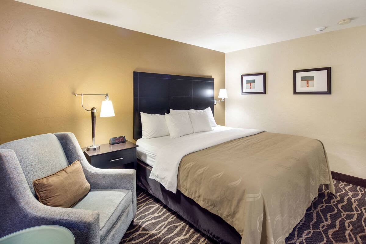 Una habitación de hotel con una cama grande, una mesita de noche, una lámpara, una silla y dos cuadros en la pared.
