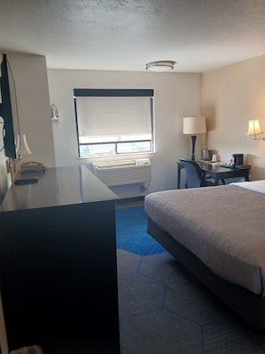 Habitación de hotel con cama, escritorio, lámpara, aire acondicionado y ventana con persianas.