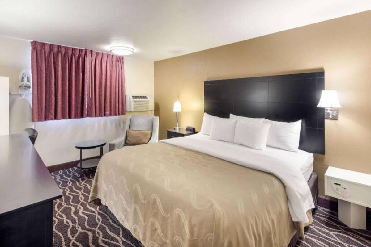 Una habitación de hotel con una cama, mesitas de noche, un escritorio, una silla, una ventana con cortinas e un equipo de aire acondicionado instalado en la pared.