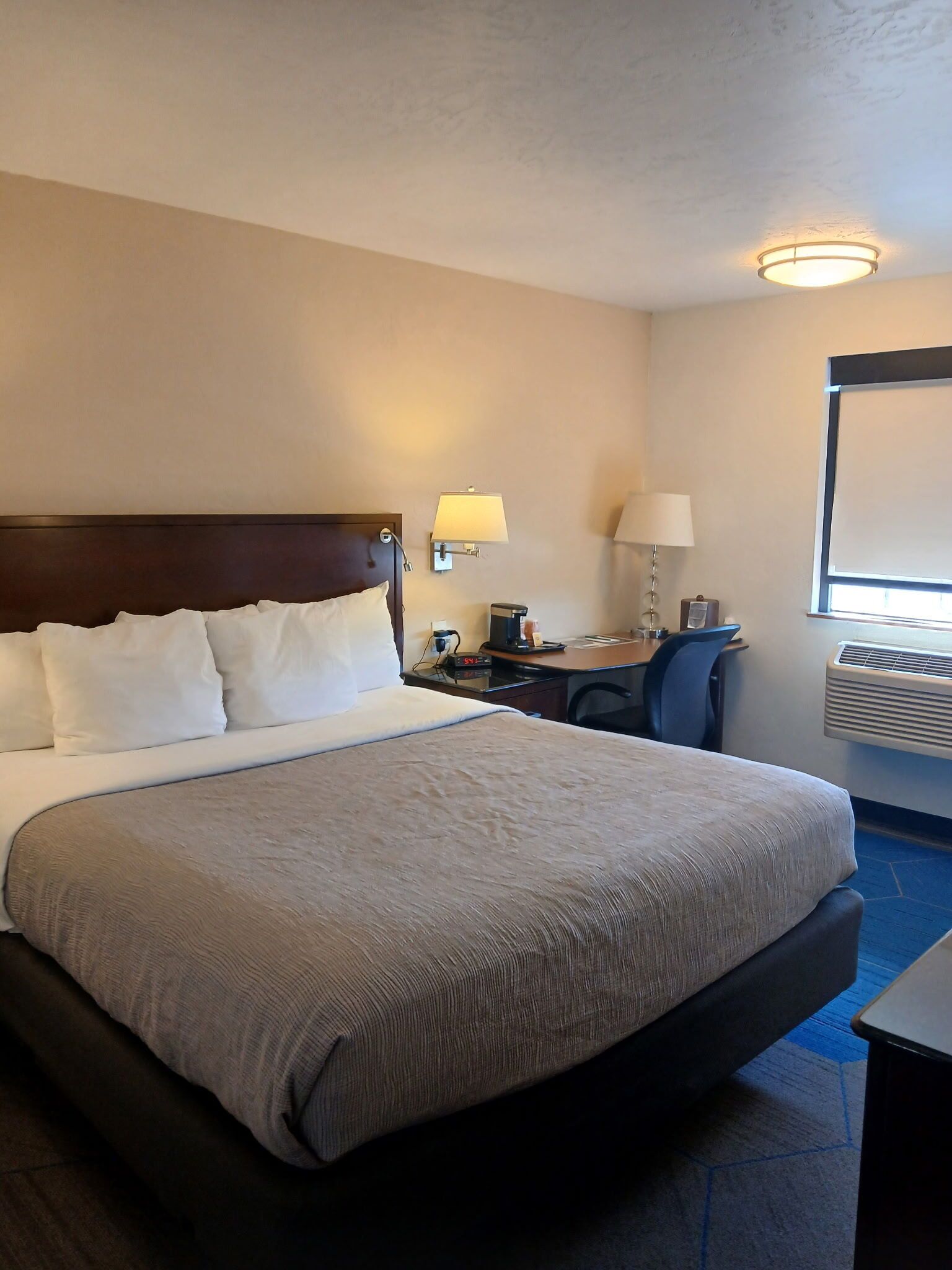 Een hotelkamer met een groot bed, een bureau, een stoel, een lamp, een klok en een airconditioningsysteem.