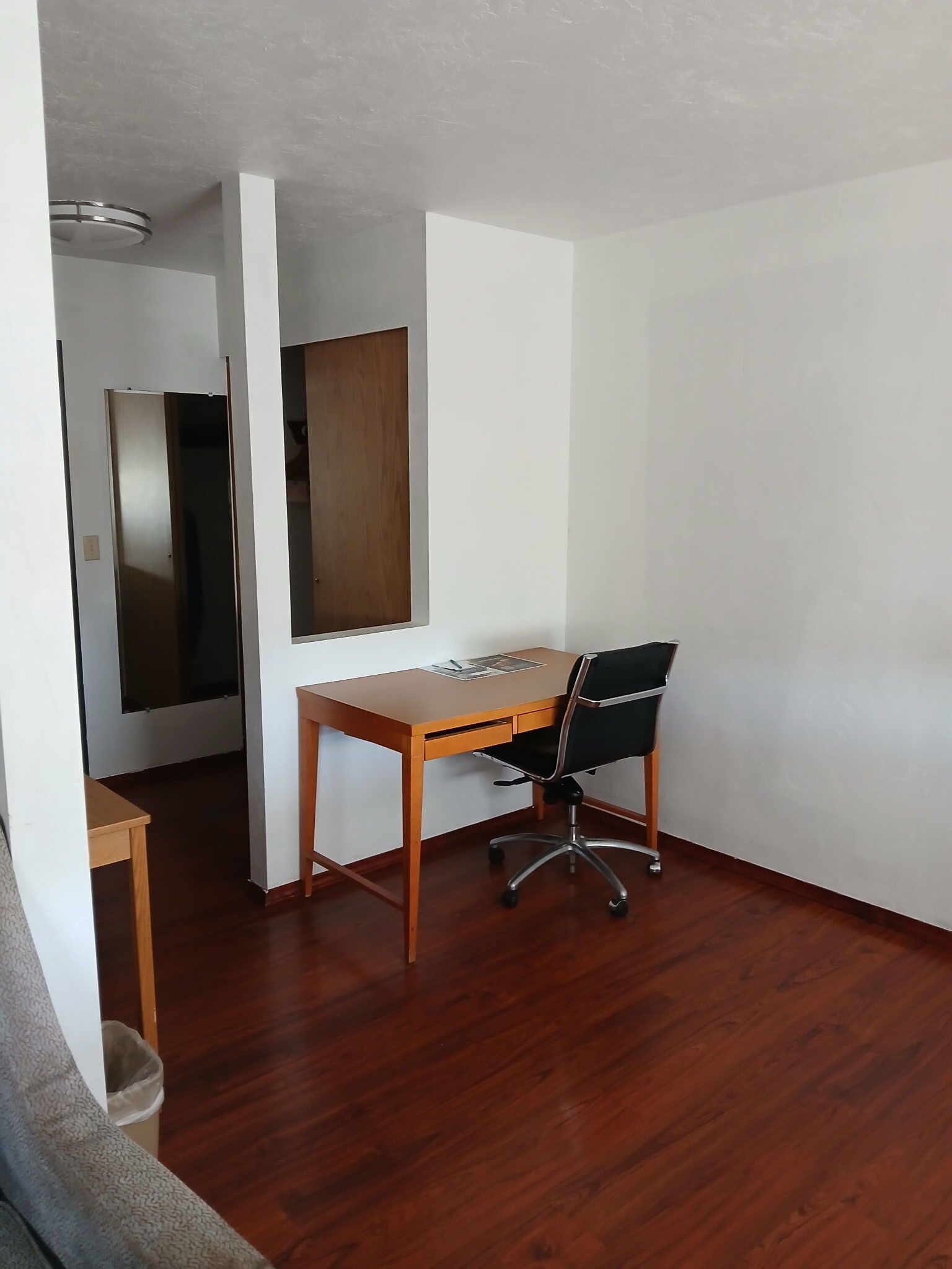 A room with a wooden desk, a black office chair, a wooden door, and a white wall.