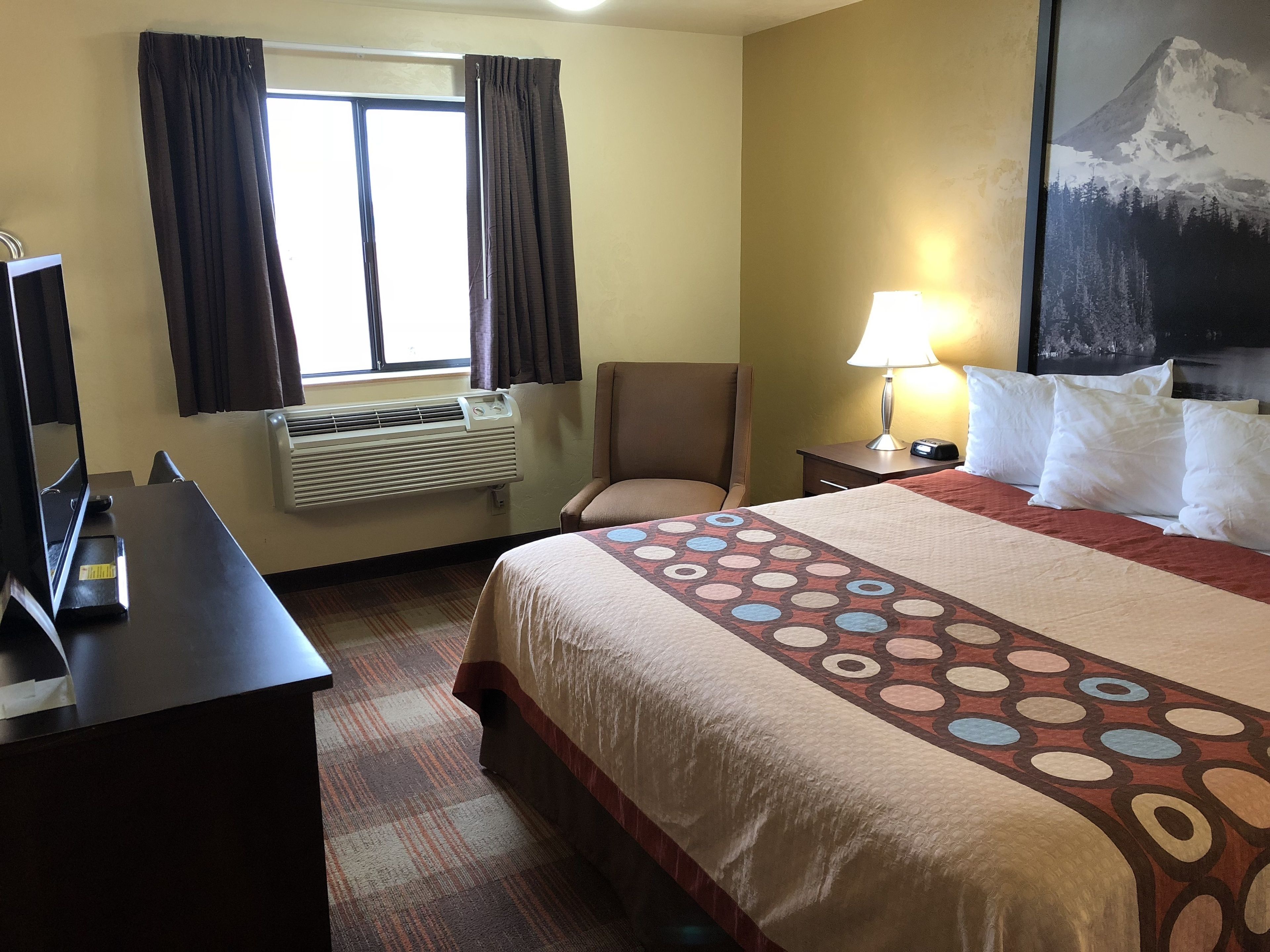 Une chambre d’hôtel avec un lit, une chaise, un bureau, une lampe et une fenêtre avec des rideaux.