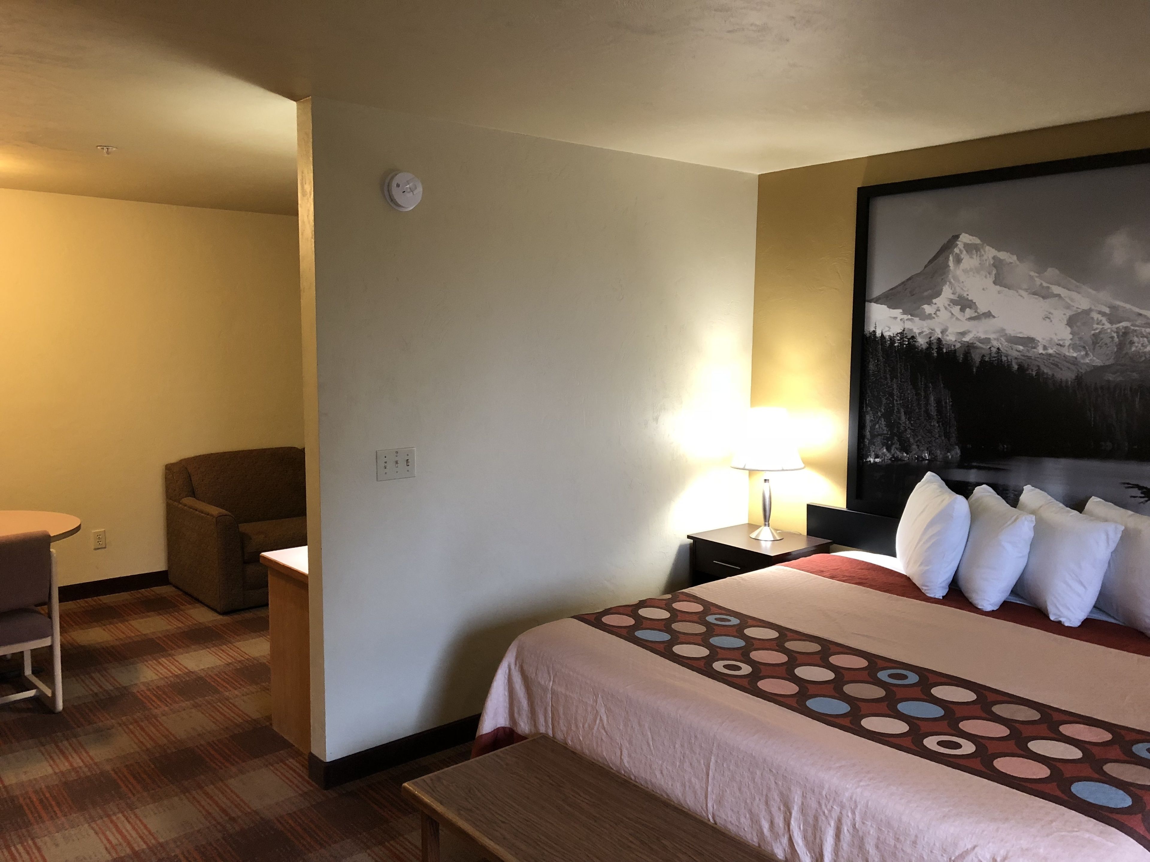 Habitación de hotel con cama, escritorio, silla, lámpara y un cuadro de una montaña.